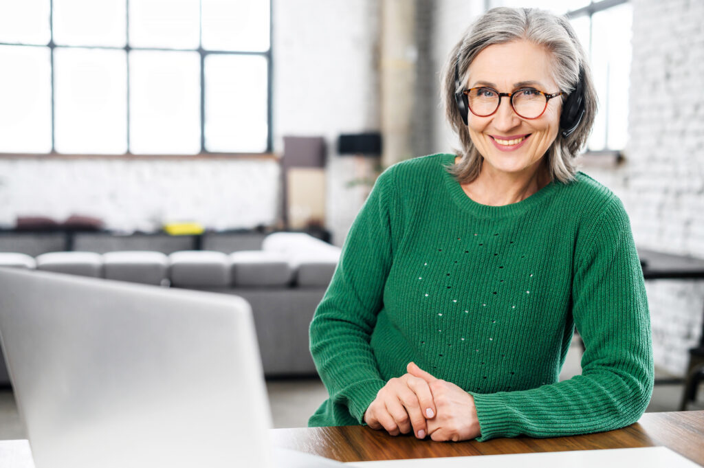 A portrait of cheerful senior woman wearing wireless headset and stylish eyeglasses, a mature female support employee sitting at the desk with a laptop and looks at the camera friendly